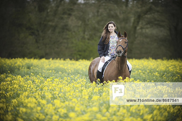 Eine Frau reitet auf einem braunen Pferd durch eine blühende gelbe Senfpflanze auf einem Feld.