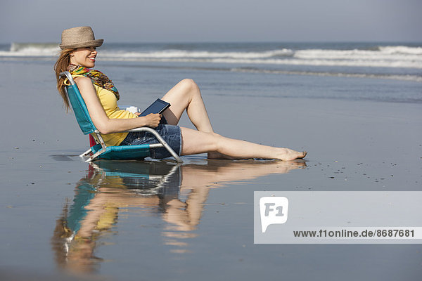 Eine Frau mit Sonnenhut und Schal am Strand der New Jersey Shore  die mit einem digitalen Tablet in der Hand sitzt.