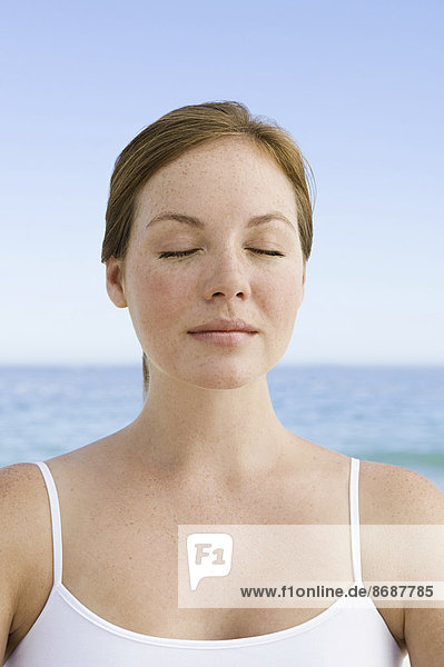 Eine junge Frau sitzt in entspannter Pose mit geschlossenen Augen am Strand.