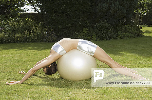 Eine junge Frau zeigt ihre Beweglichkeit  lehnt sich rückwärts über einen Übungsball  mit Händen und Füßen auf Gras.