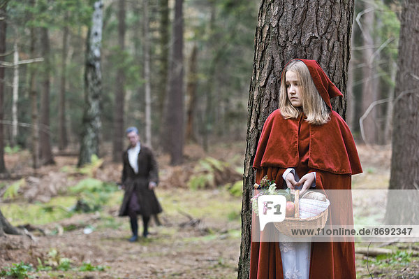 Girl masquerade as Red Riding Hood hiding behind a tree in the wood