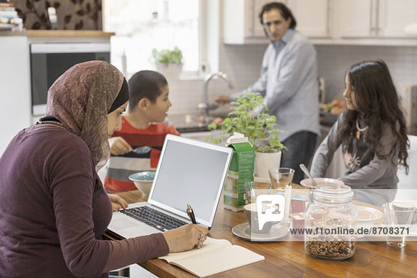Muslim woman working on laptop with family having breakfast in kitchen