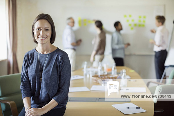 Portrait of confident businesswoman sitting on conference table with colleagues discussing in background