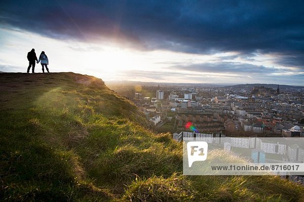 A young couple hold hands in front of the view of the City of Edinburgh from Salisbury Crags