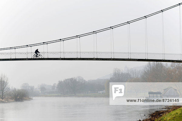 Radfahrer beim Überqueren einer Hängebrücke  Wassermühle im Fluss Weser  Minden  Nordrhein-Westfalen  Deutschland