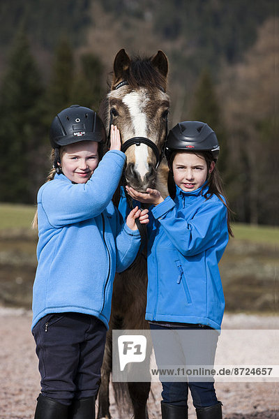 Two girls wearing riding helmets standing beside a pony  dun  with a bridle  Tyrol  Austria