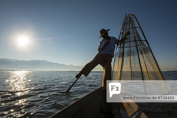 Fisherman in the morning light leg rower with a traditional basket on the canoe at sunrise Inle Lake Shan State Myanmar
