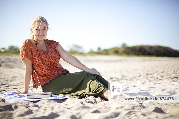 Portrait of Young Woman with Book  Looking at Camera and Sitting on Beach  Palm Beach Gardens  Palm Beach County  Florida  USA