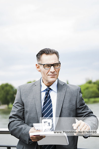 Portrait of mature businessman standing on bridge holding documents  wearing horn-rimmed eyeglasses  Mannheim  Germany