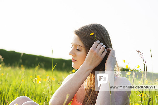 Young woman sitting in field placing flower in hair  Germany