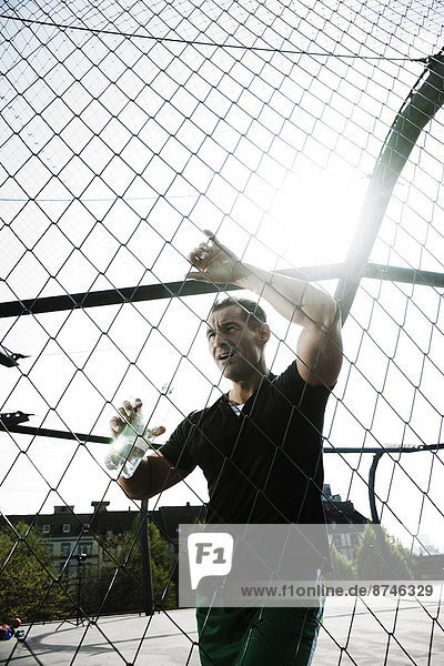 Mature man leaning against chain-link fence on outdoor basketball court  holding bottle of water  Germany