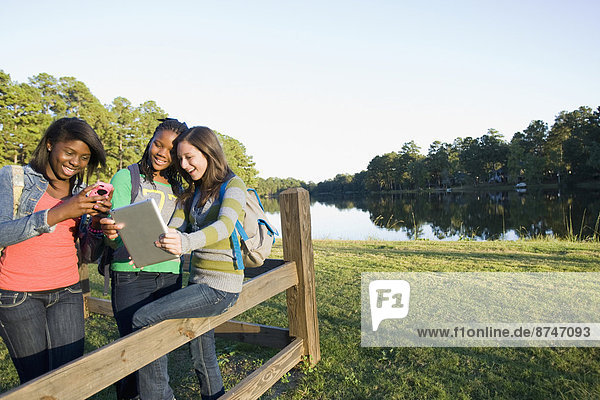 Pre-teen girls sitting on fence  looking at tablet computer and cellphone  outdoors
