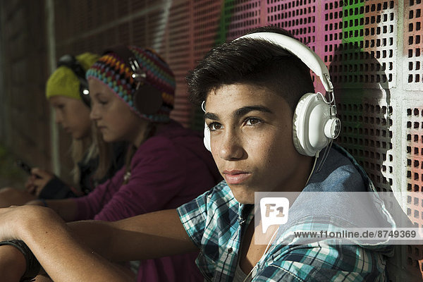 Close-up portrait of boy sitting next to wall  wearing headphones and listening to music  other children in background  Germany