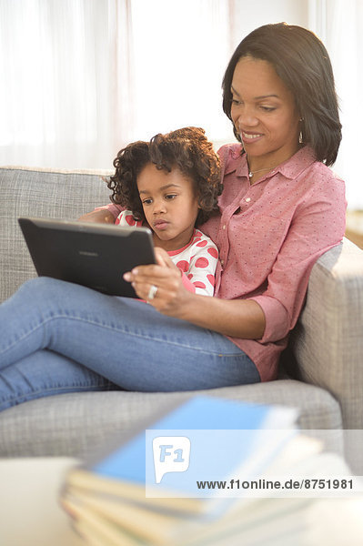 Black mother and daughter using digital tablet on sofa
