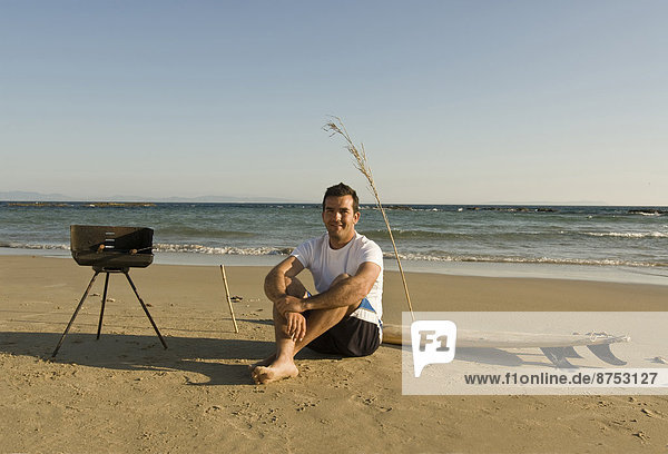 man sitting on beach with surfboard and barbecue