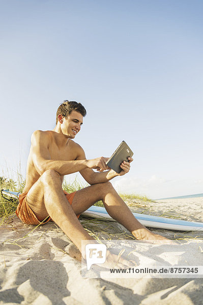 Young man sitting on beach with surfboard and digital tablet