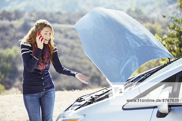 Frustrated woman talking on cell phone with automobile hood raised at roadside