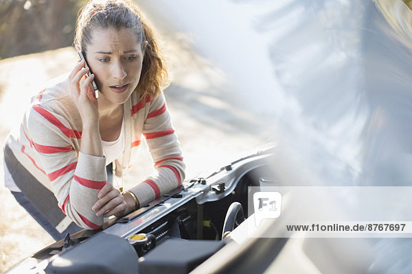 Frustrated woman talking on cell phone and looking at car engine at roadside