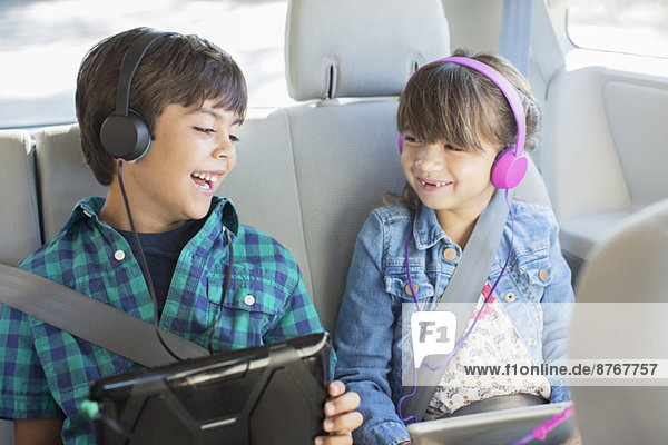 Glückliche Geschwister mit Kopfhörern und digitalen Tabletts auf dem Rücksitz des Autos
