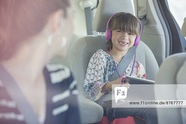 Mutter dreht sich um und lächelt die Tochter mit Kopfhörer und digitalem Tablett auf dem Rücksitz des Autos an.