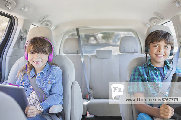 Portrait von glücklichen Geschwistern mit Kopfhörern und digitalen Tabletts auf dem Rücksitz des Autos
