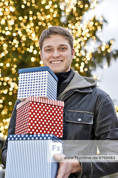 Junger Mann mit drei Geschenkboxen vor dem beleuchteten Weihnachtsbaum