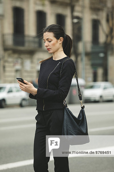 Spain  Catalunya  Barcelona  young black dressed businesswoman with smartphone in front of a street