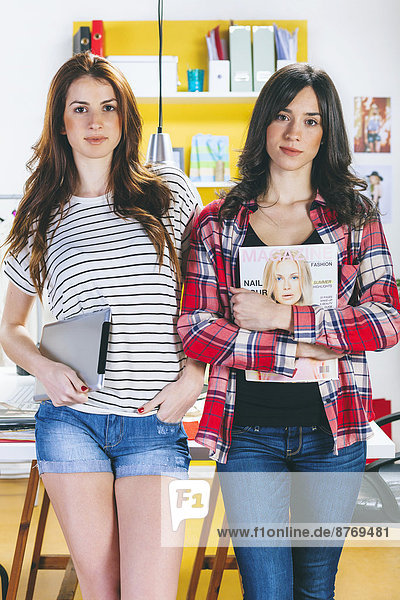 Portrait of two female fashion bloggers with digital tablet and magazine at office