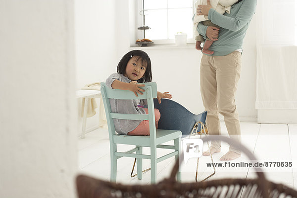 Kleines asiatisches Mädchen auf einem Stuhl sitzend  Mutter und Schwester im Hintergrund