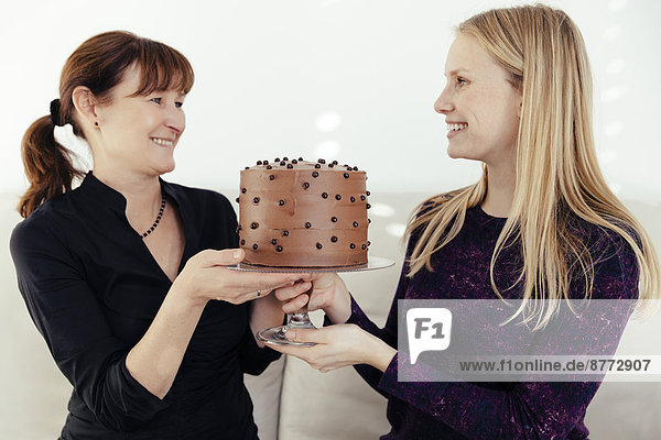 Two smiling women exchanging cake stand with chocolate cake