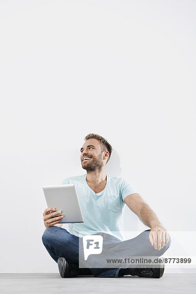 Porträt eines jungen Mannes  der auf dem Boden sitzt und einen Tablet-Computer hält.