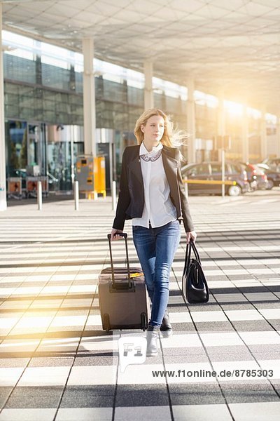 Junge Frau geht vor dem Flughafen und zieht einen Rollkoffer