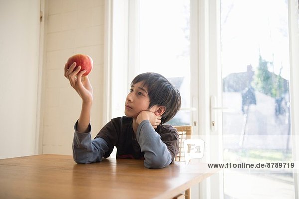 Junge sitzt am Tisch und hält einen Apfel vor sich.