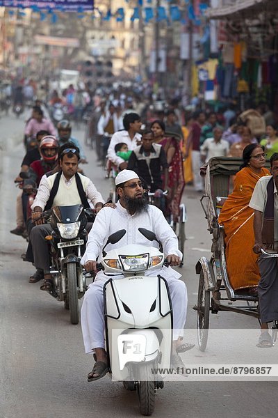 Muslim man wearing topi cap and white outfit driving motor scooter in street scene in city of Varanasi  Benares  Northern India
