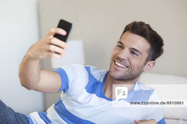 Junger Mann liegt auf dem Bett und nimmt Selfie mit dem Smartphone.
