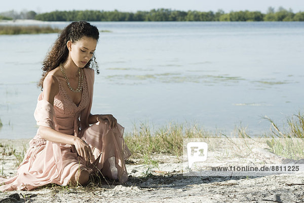 Junge Frau im Kleid am See sitzend  mit Stock in Sand gezeichnet