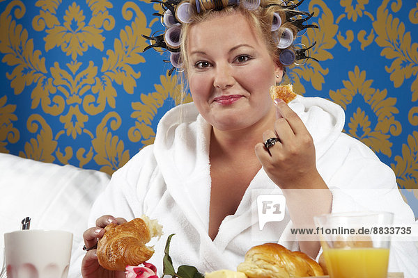 Portrait of woman with curlers and white bathrobe having breakfast in bed