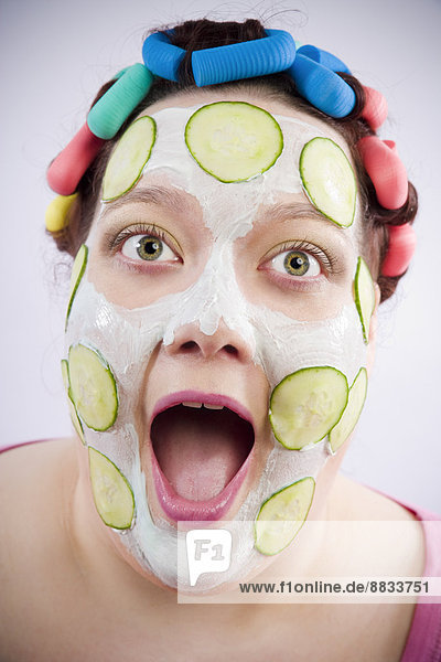 Porträt der schreienden Frau mit Lockenwickler und Schönheitsmaske mit Gurkenscheiben