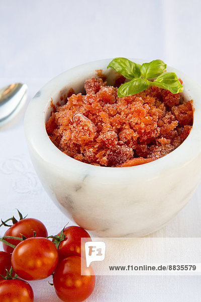 Marmorschale mit Tomatengranita  garniert mit Basilikumblättern und Kirschtomaten auf weißem Tuch