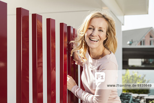 Lächelnde Frau steht am roten Zaun des Wohnhauses