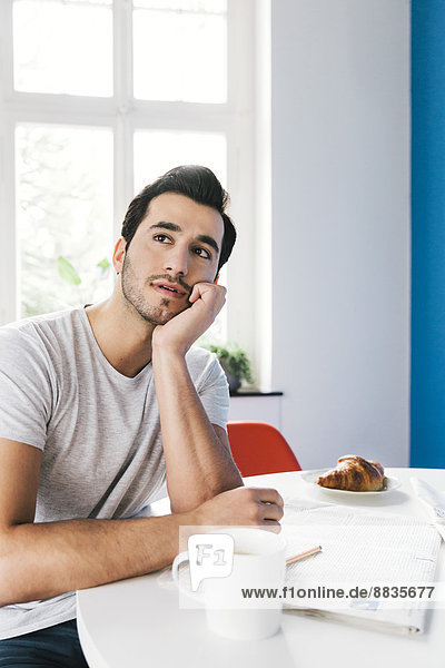 Portrait of pondering young man sitting at breakfast table with newspaper