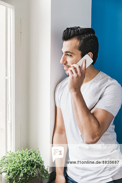 Porträt eines jungen Mannes  der mit einem Smartphone in seiner Küche telefoniert.