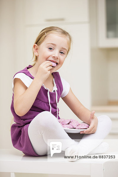 Porträt eines lächelnden kleinen Mädchens  das auf einem Tisch sitzt und rosa Kekse isst.