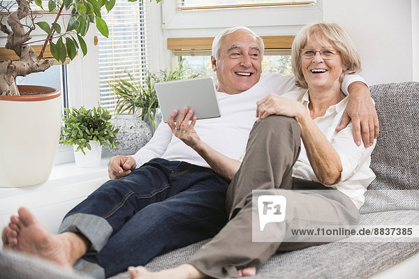 Seniorenpaar mit digitalem Tablett nebeneinander auf Sofa im Wohnzimmer