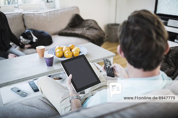 Mann hält digitales Tablett  während er zu Hause auf dem Sofa sitzt.