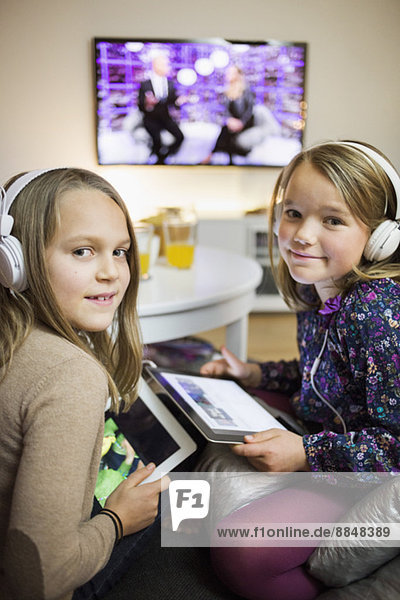 Porträt von lächelnden Schwestern beim Musikhören auf digitalen Tabletts im Wohnzimmer