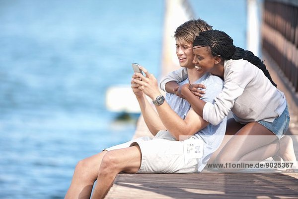 Junges Paar sitzt auf einem Steg und schaut auf sein Handy