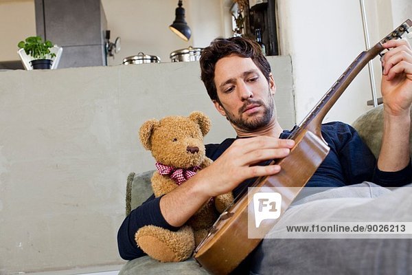 Mittlerer Erwachsener Mann der auf dem Sofa liegt und Gitarre spielt.
