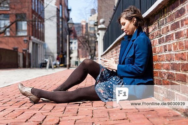 Young woman sitting against a brick wall using smartphone