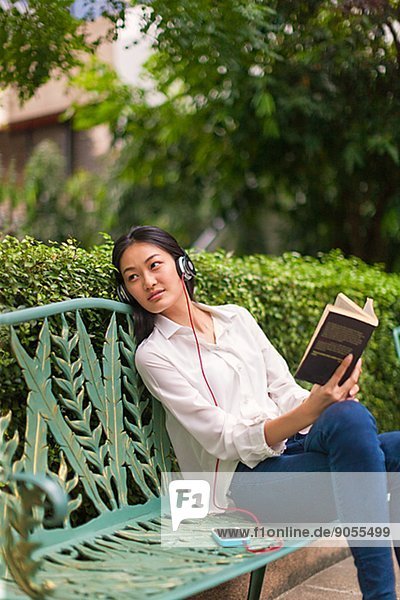 Young woman with headphones and book on park bench  Bangkok  Thailand